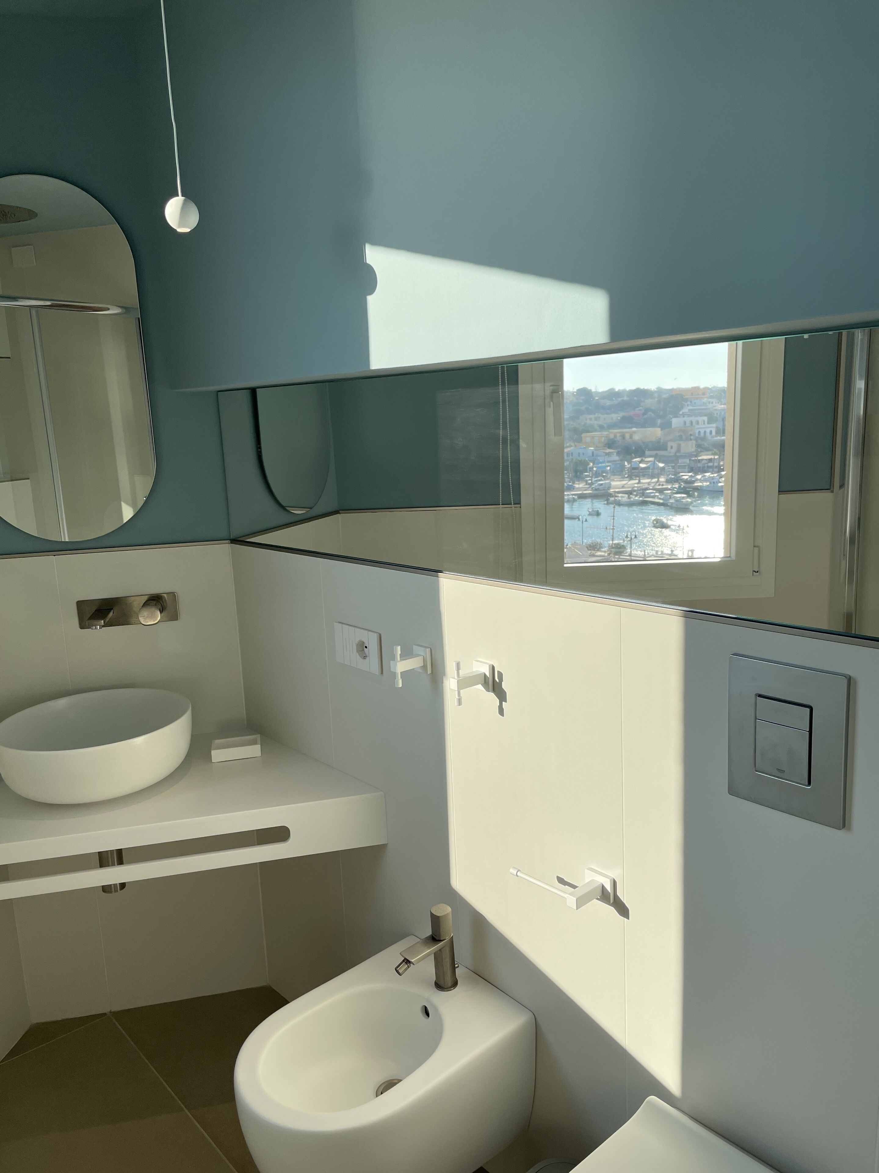 A modern bathroom with a large mirror, a round sink, a bidet, and a window offering a view of a harbor.