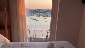 Ein Hotelzimmer mit einem Bett, zwei mit Schrift bedruckte Handtücher, einem Balkon mit Tisch und Stühlen und Blick auf Boote auf dem Wasser.