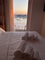 Habitación de hotel con cama, dos toallas enrolladas con texto, balcón con mesa y sillas, y vista a barcos en el agua.