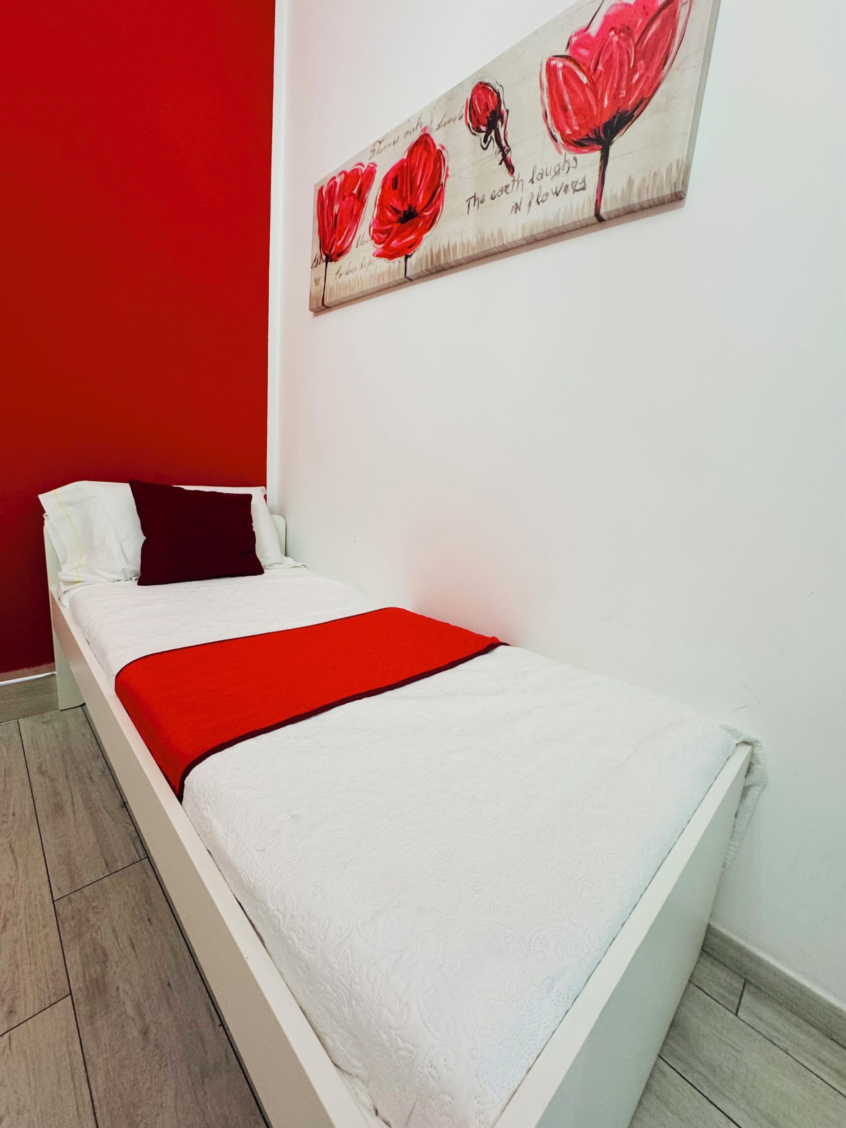 Un llit individual amb matalàs blanc, llençol vermell i almoines en una habitació amb una paret vermella d'accent i un quadre de flors vermelles.