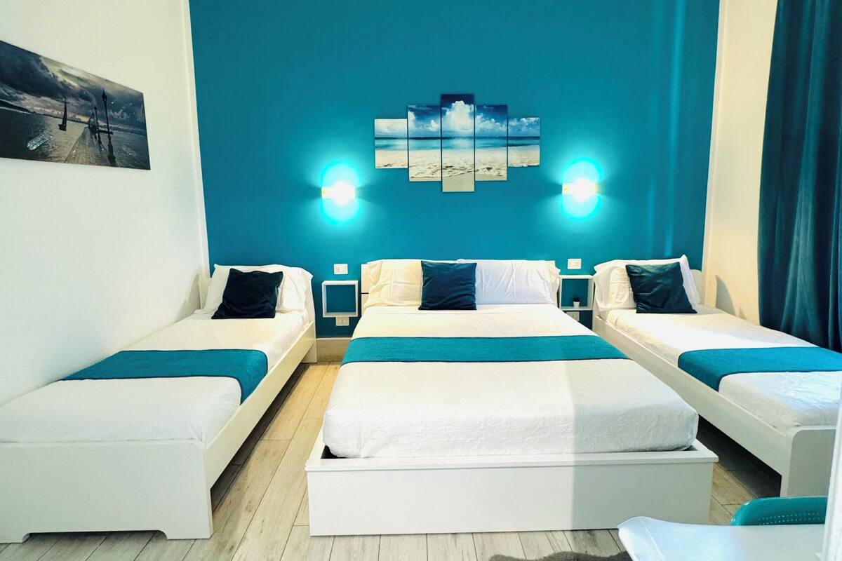 Ein Hotelzimmer mit zwei Einzelbetten, einem Doppelbett, einer Deckenleuchte, einem Wandbild und einem Nachttisch mit Lampe.