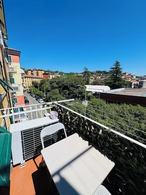 A balcony with a table and chairs, overlooking a street with parked cars and buildings.