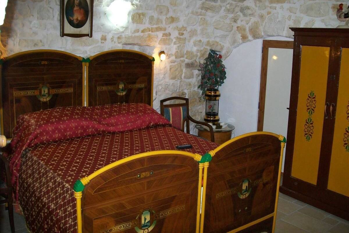 Une chambre avec un mur en pierre, un lit en bois recouvert d’une courtepointe rouge, une armoire en bois et un tableau encadré accroché au mur.