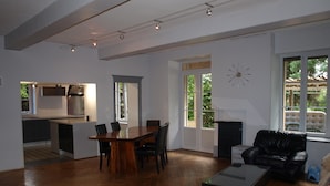A spacious living area with a wooden herringbone floor, a dining table with chairs, a black leather sofa, and a coffee table. There is a kitchen in the background with a stainless steel range hood. - Chambre d'hôte Calme et Volupté (Maxilly-sur-Saône)