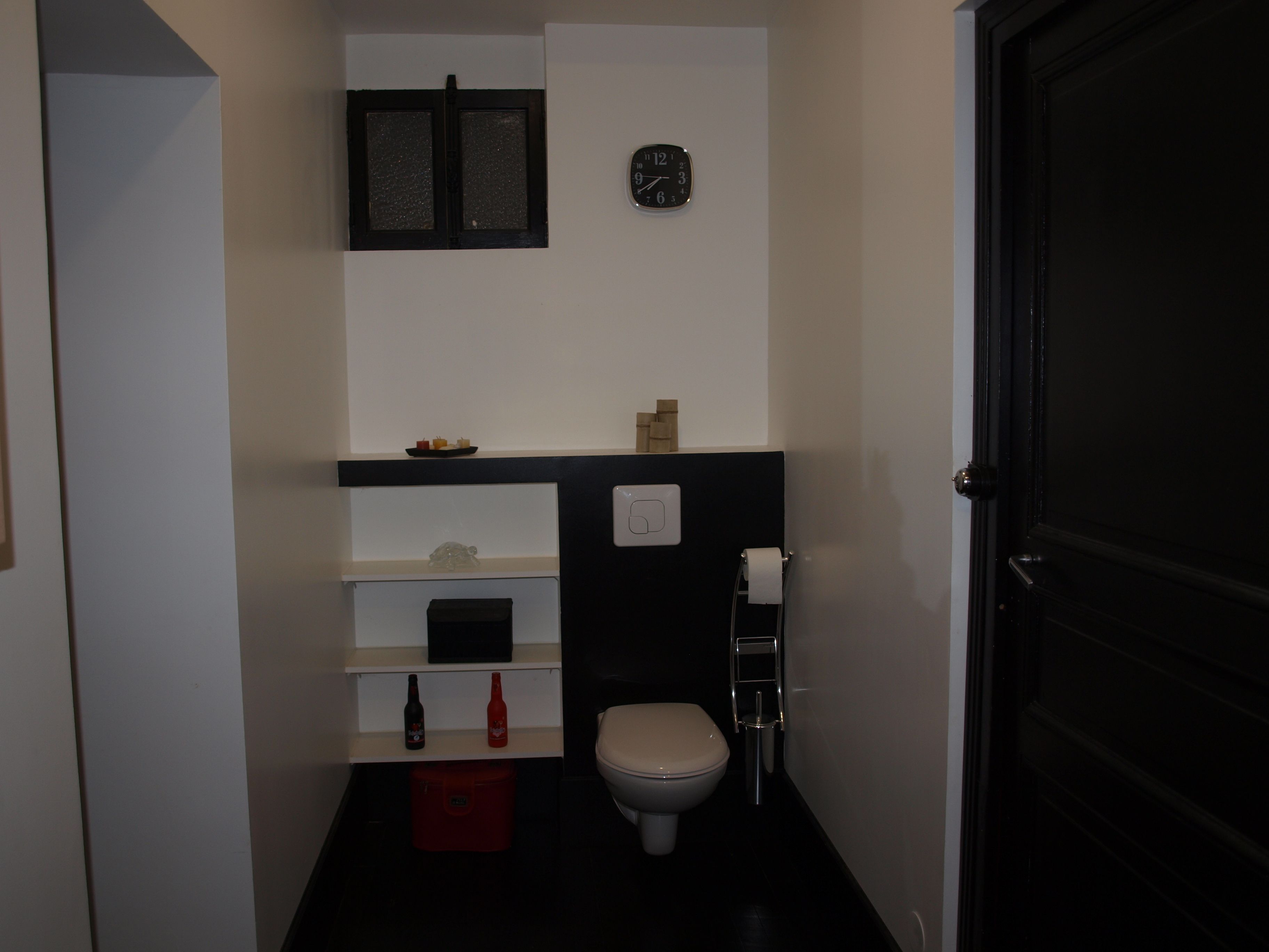 Casa de banho pequena com sanita, prateleira com artigos de higiene, relógio e porta.