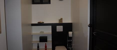 Casa de banho pequena com sanita, prateleira com artigos de higiene, relógio e porta.