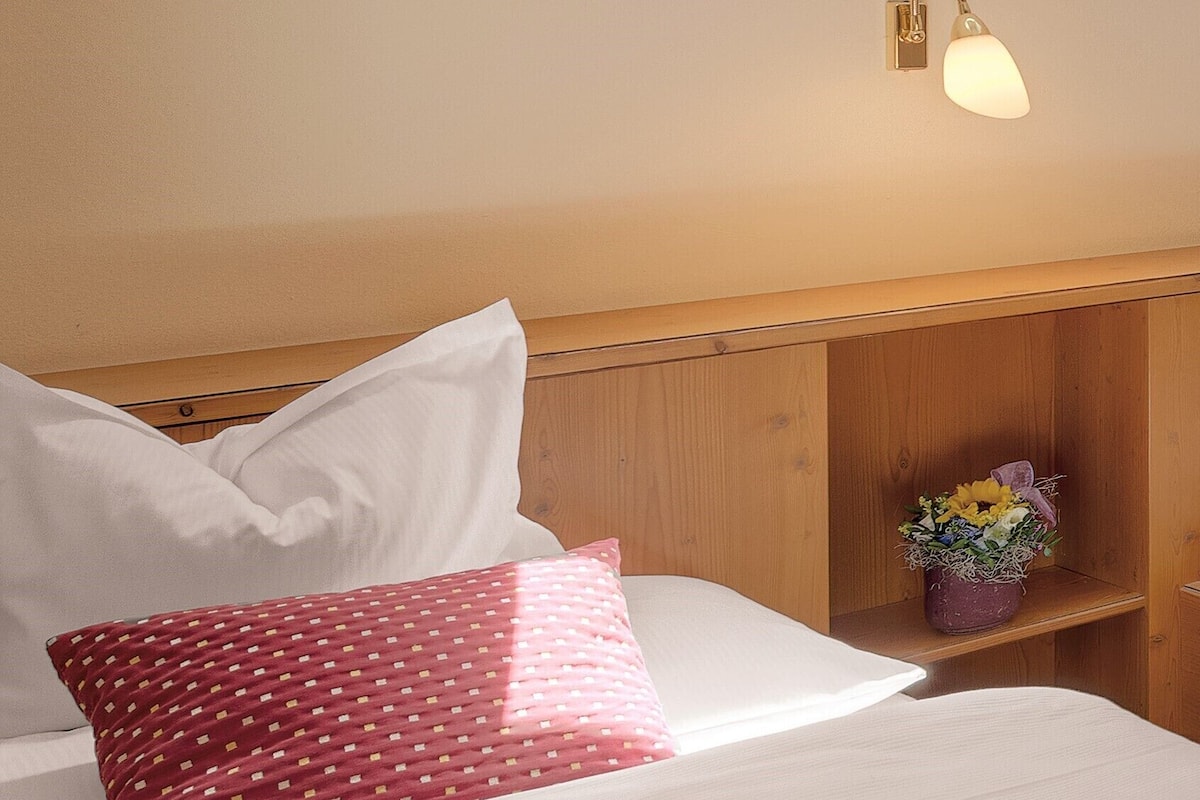 Una cama bien hecha con almohadas blancas y rojas, un cabecero de madera y una pequeña estantería con un arreglo floral.