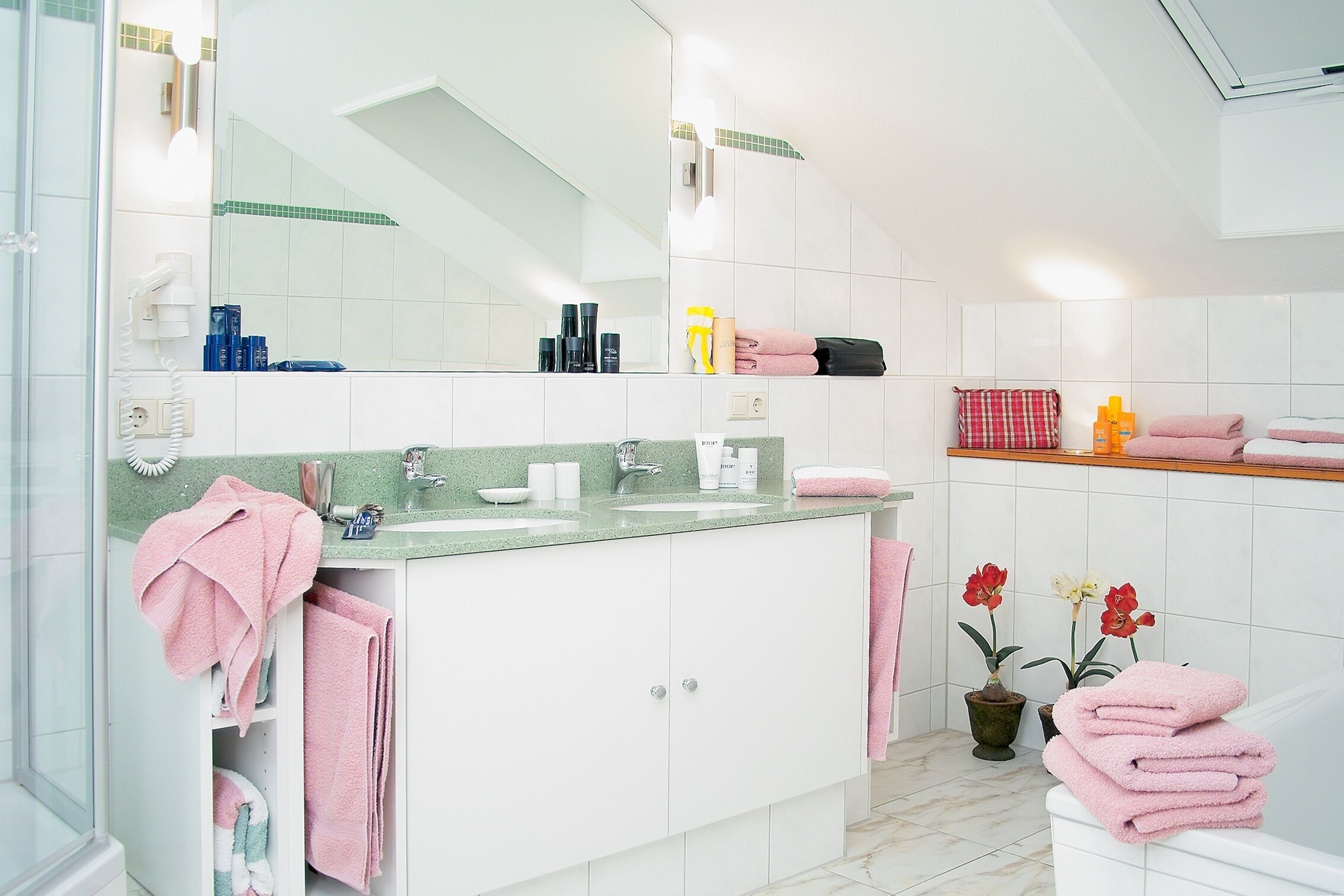 A modern bathroom with a white vanity, a large mirror, and a sink. There are towels and toiletries on the counter, and two potted plants on the floor.