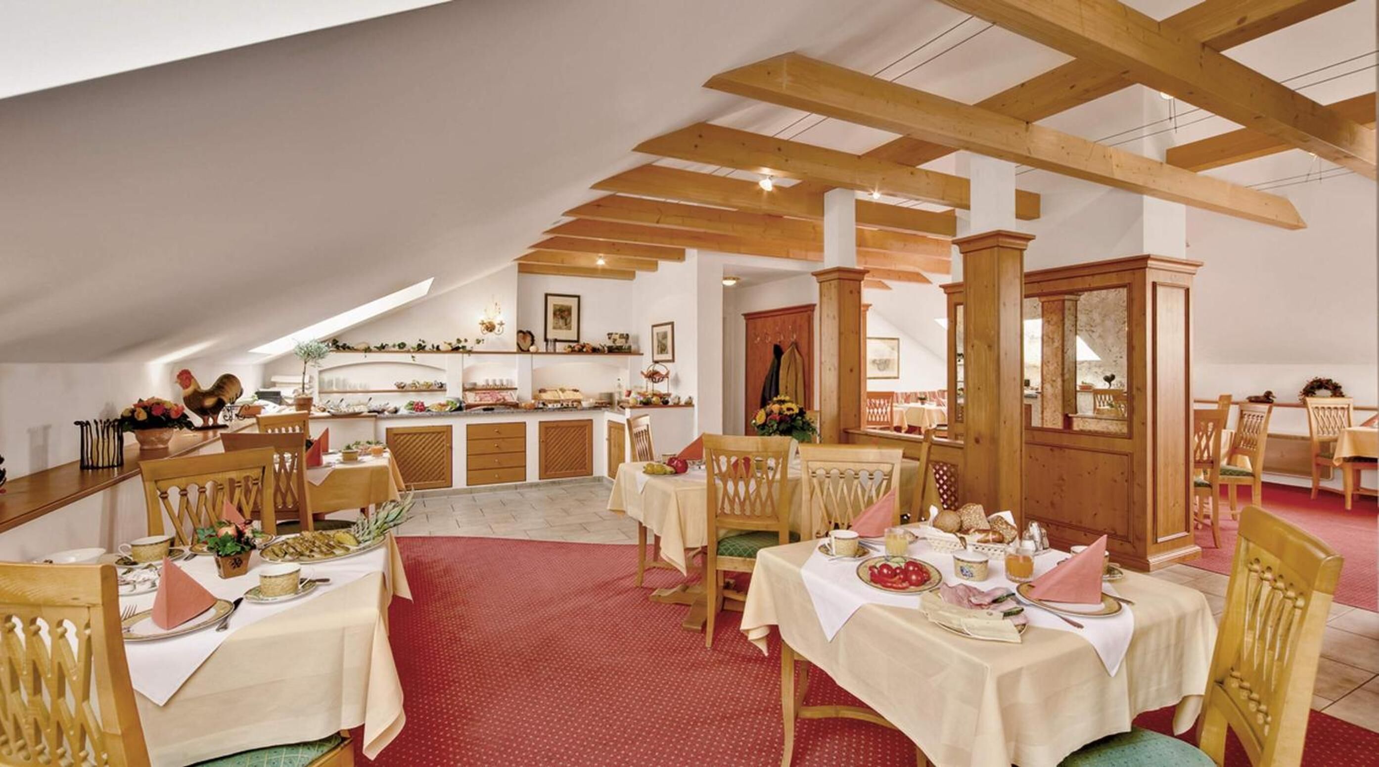 Uma sala de jantar com vigas de madeira, mesas cobertas com toalhas brancas e um tapete vermelho.