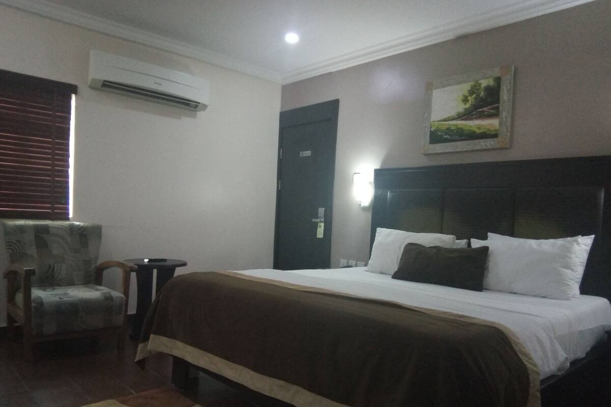A hotel room with a bed, a chair, an air conditioning unit, a door, and a wall-mounted light fixture.