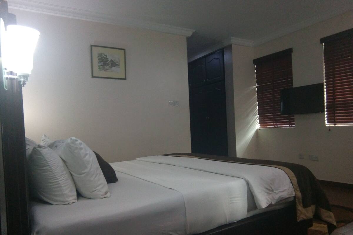 A bedroom with a bed, pillows, a bedside table, a lamp, a window with blinds, and a wall-mounted TV.