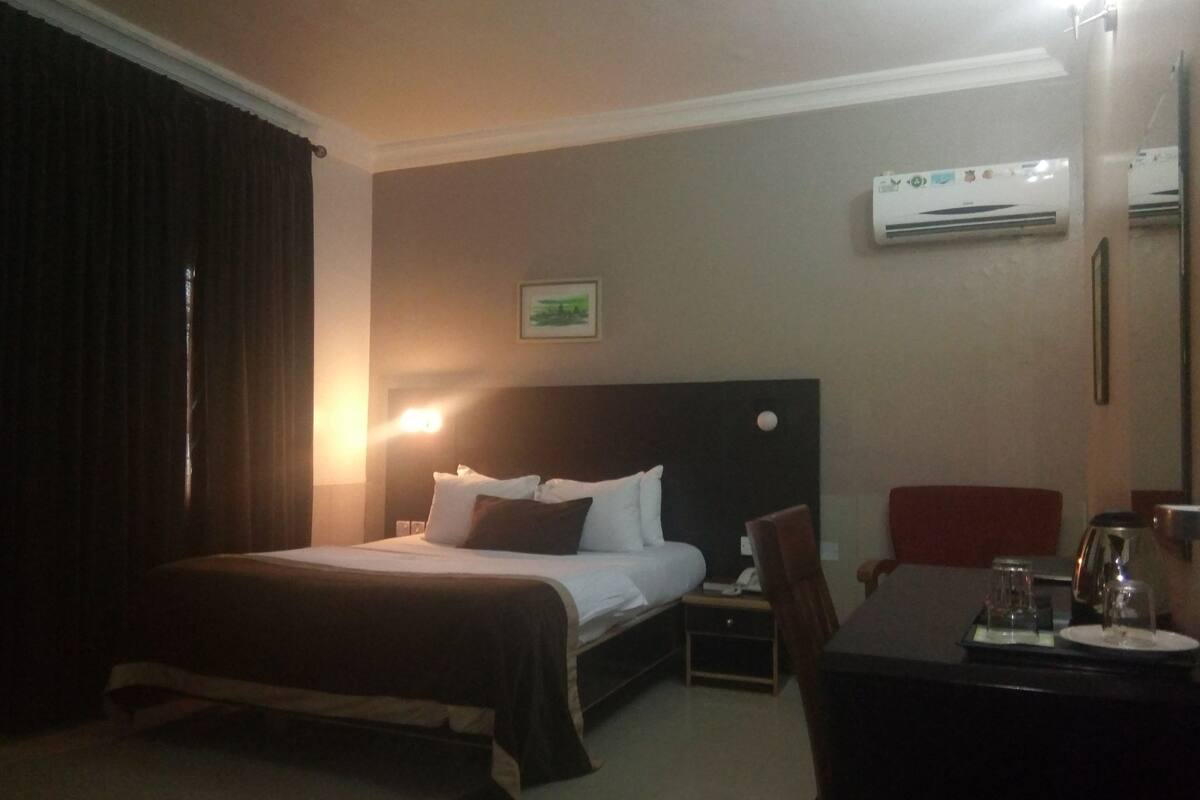A hotel room with a bed, bedside table, chair, and an air conditioning unit.
