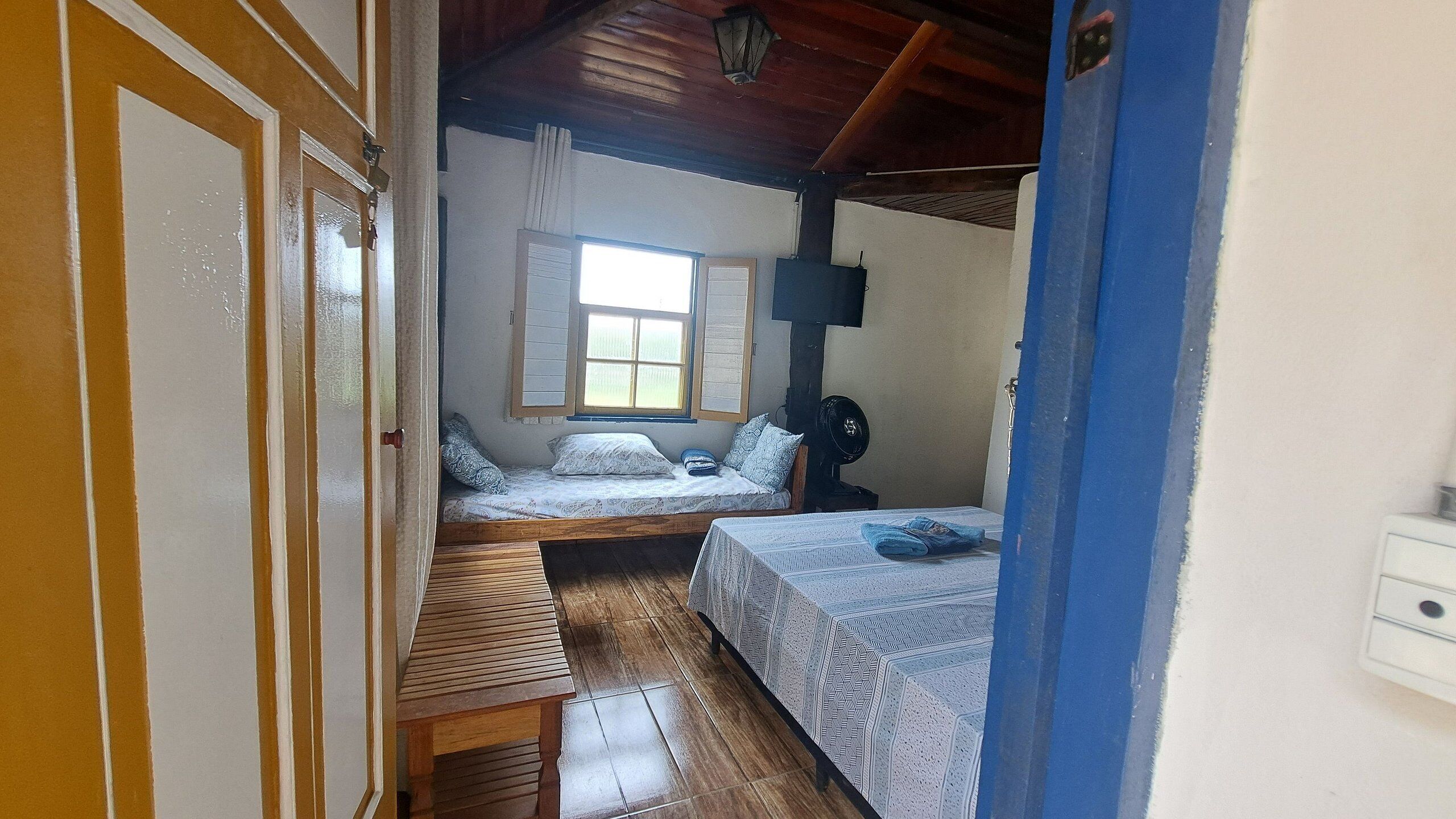 A narrow room with two beds, a wooden bench, a window, and a wall-mounted fan.