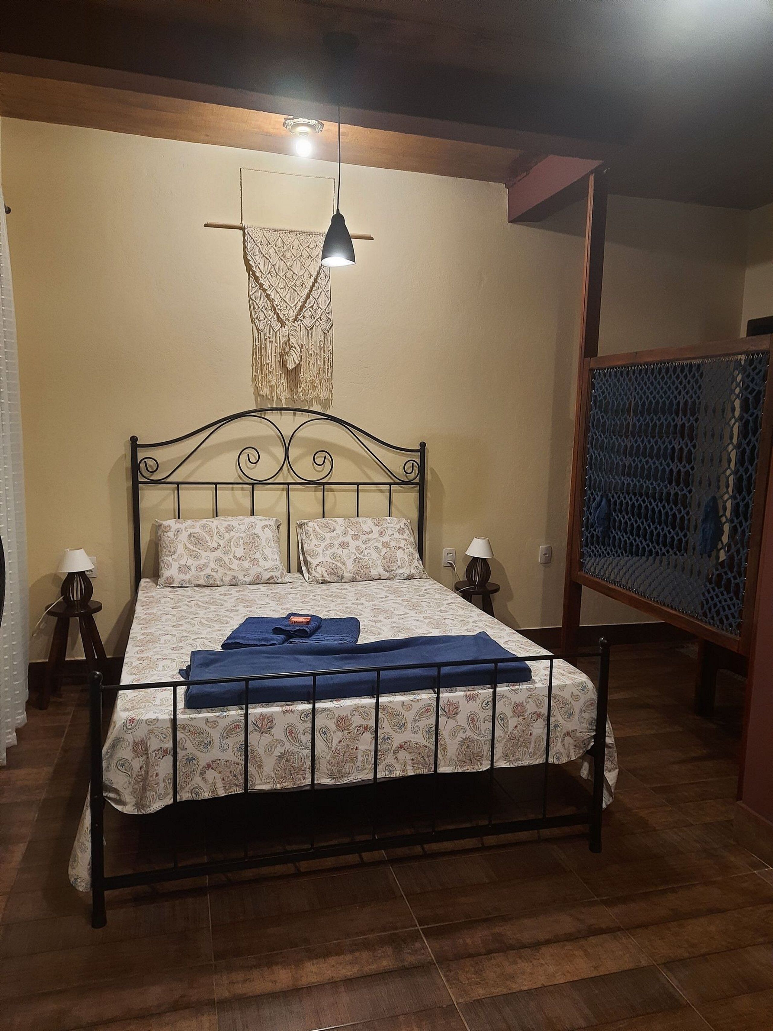 Uma cama com estrutura de metal, colcha estampada e dois travesseiros. Há uma decoração em macramê pendurada acima da cama.