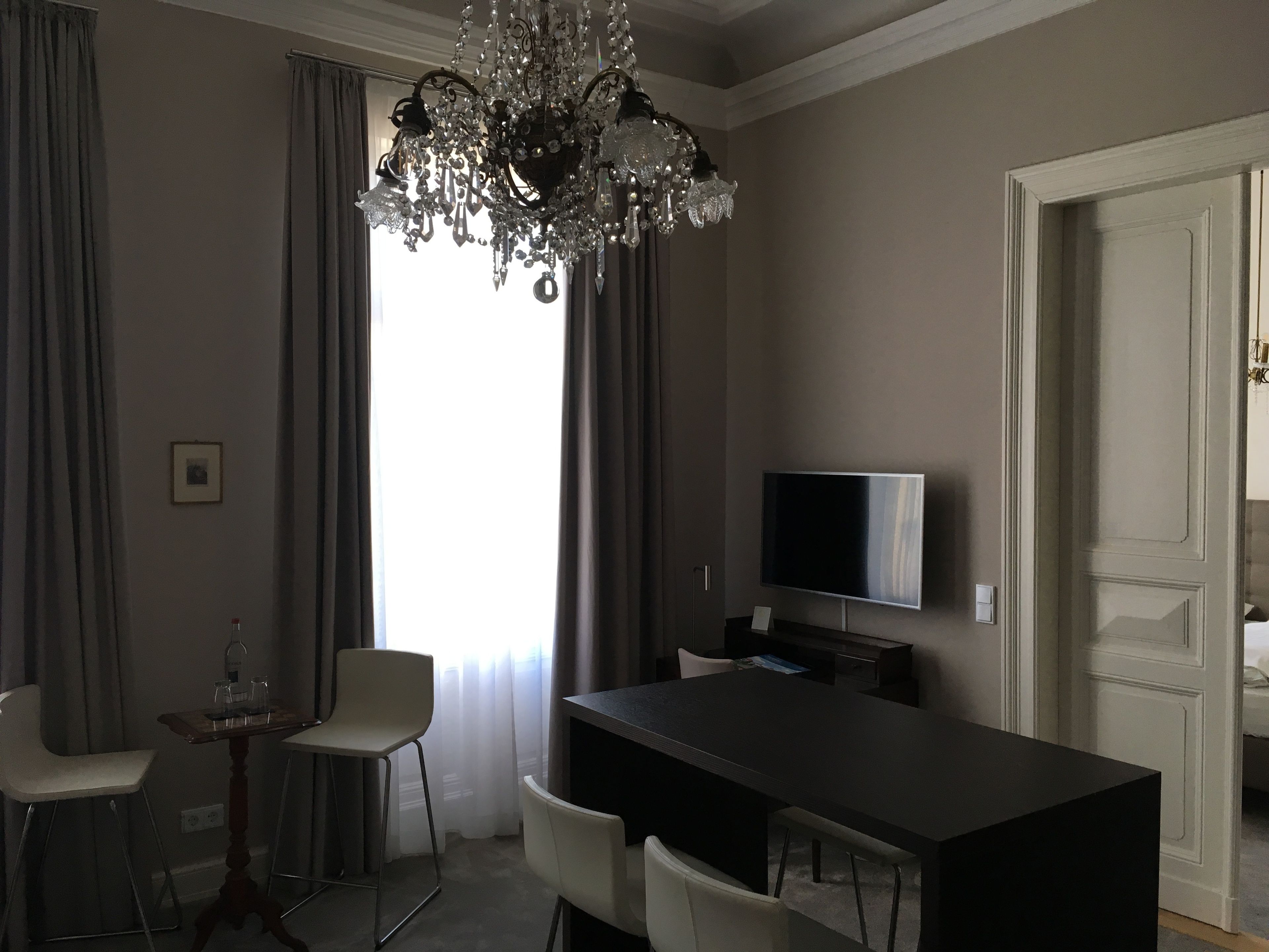 Ein modernes Hotelzimmer mit großem Fenster, einer Kristallleuchtre, einem Flachbildfernseher und einem Essbereich mit Tisch und Stühlen.
