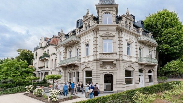 Ein historisches Gebäude mit einem zentralen Turm, zahlreichen Fenstern und einem Gartenbereich mit Besuchern.