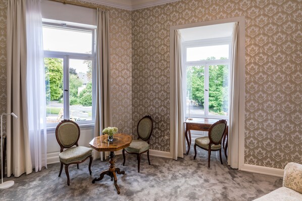 A room with two large windows, patterned wallpaper, and a round wooden table with chairs.