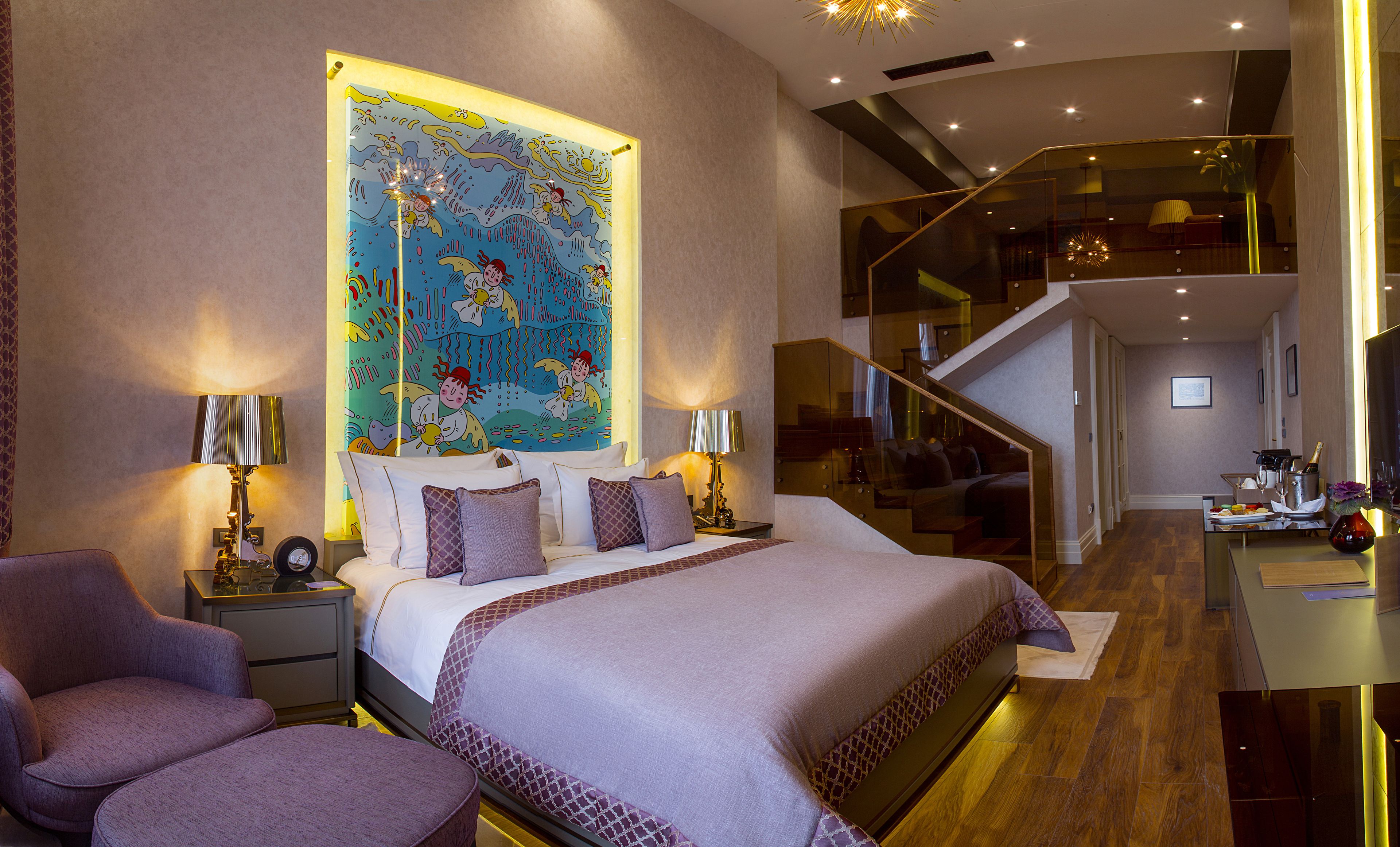 A modern hotel room with a large bed, two bedside tables with lamps, a chair, a large window with a colorful mural, and a staircase leading to an upper level.
