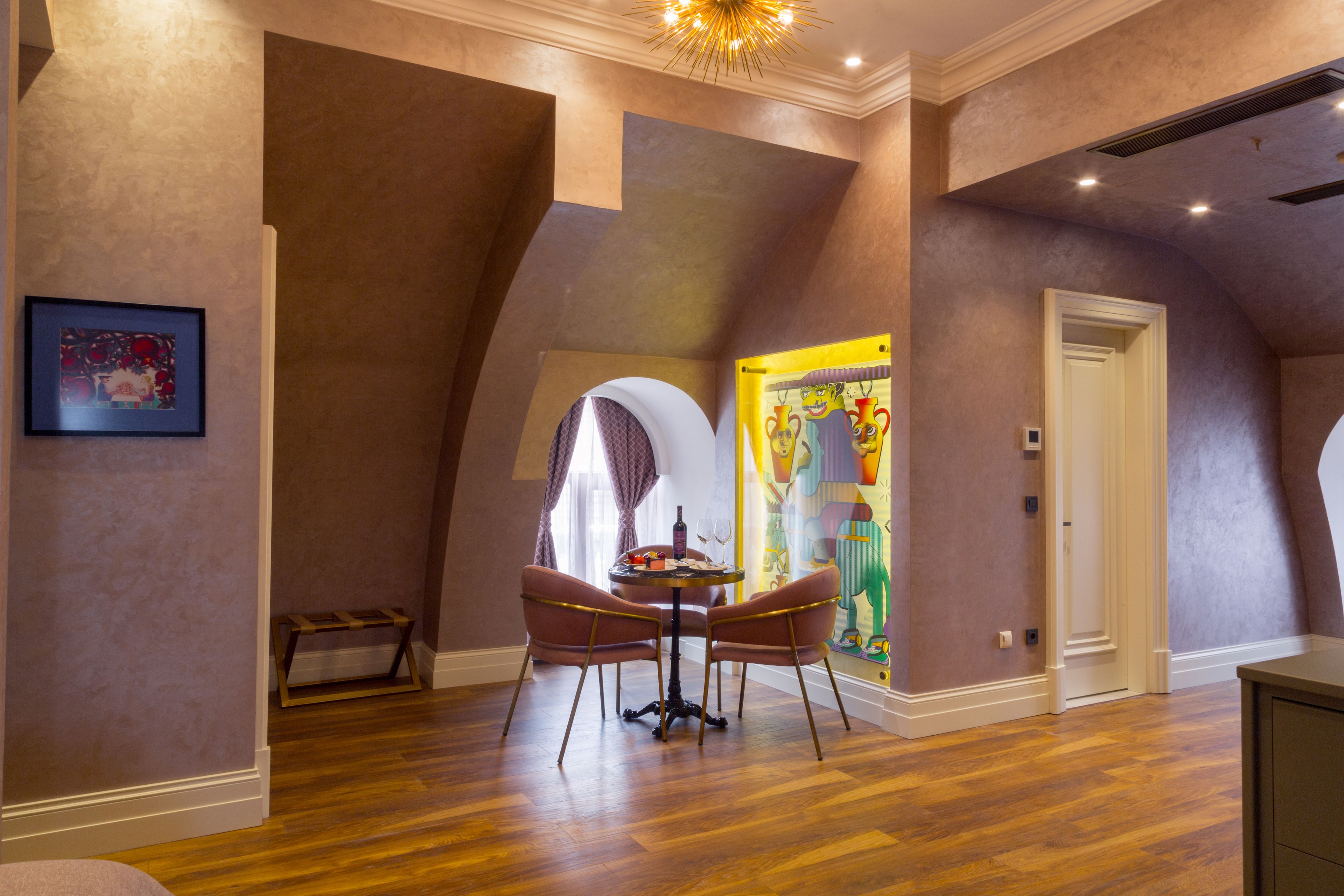 A room with a wooden floor, a round table with chairs, a door, and a colorful painting on the wall.
