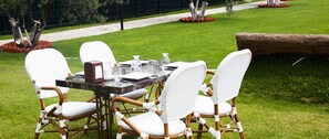 A dining table with white chairs and a glass top, set on a grassy lawn with a stone wall in the background. - Assos Altin Hotel (Ayvacik)