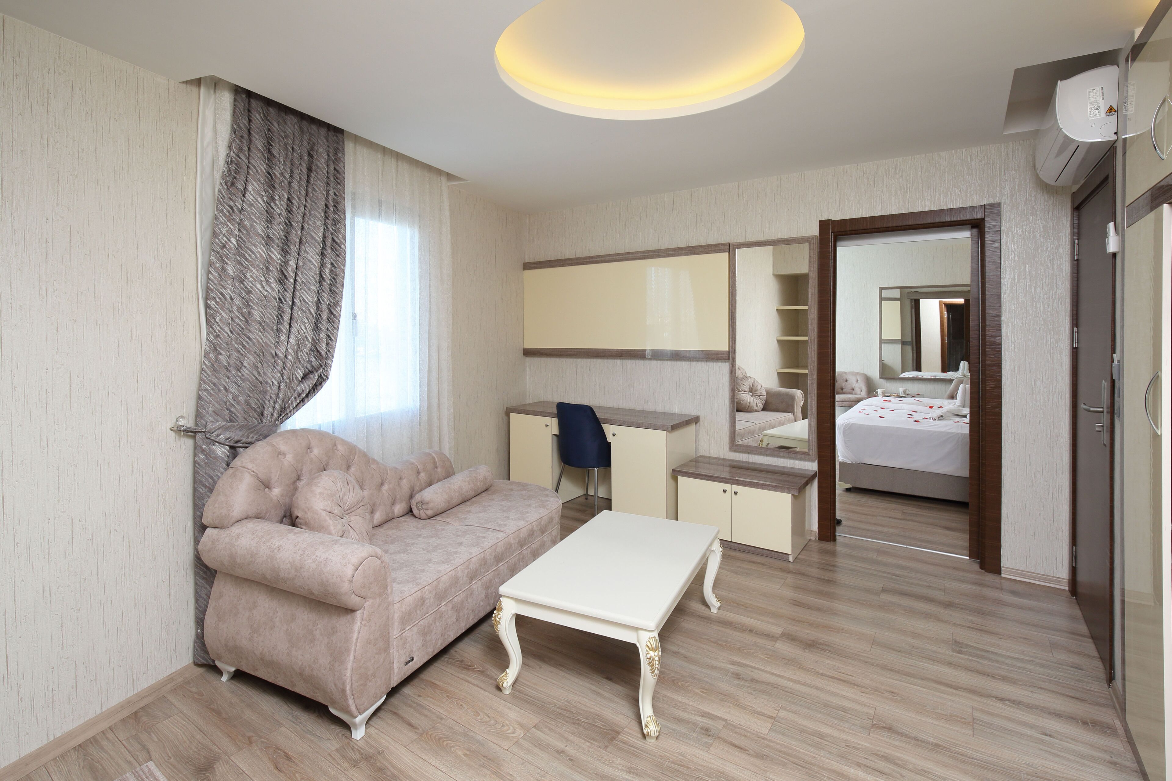 A modern hotel room with a beige sofa, a white coffee table, a desk with a chair, a large mirror, and a bed with a white duvet.