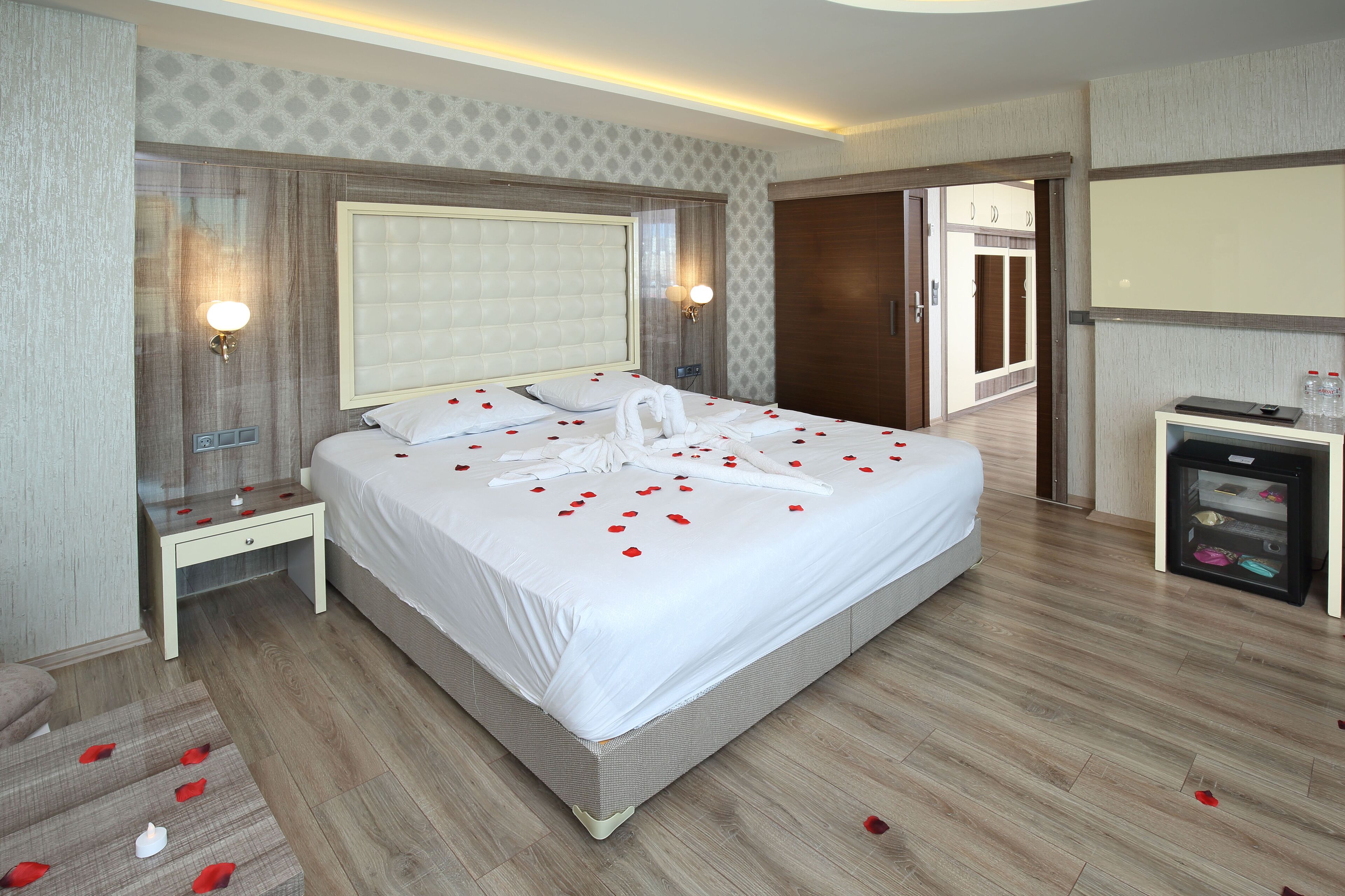 A neatly made bed with rose petals and a towel, a bedside table, and a small refrigerator.
