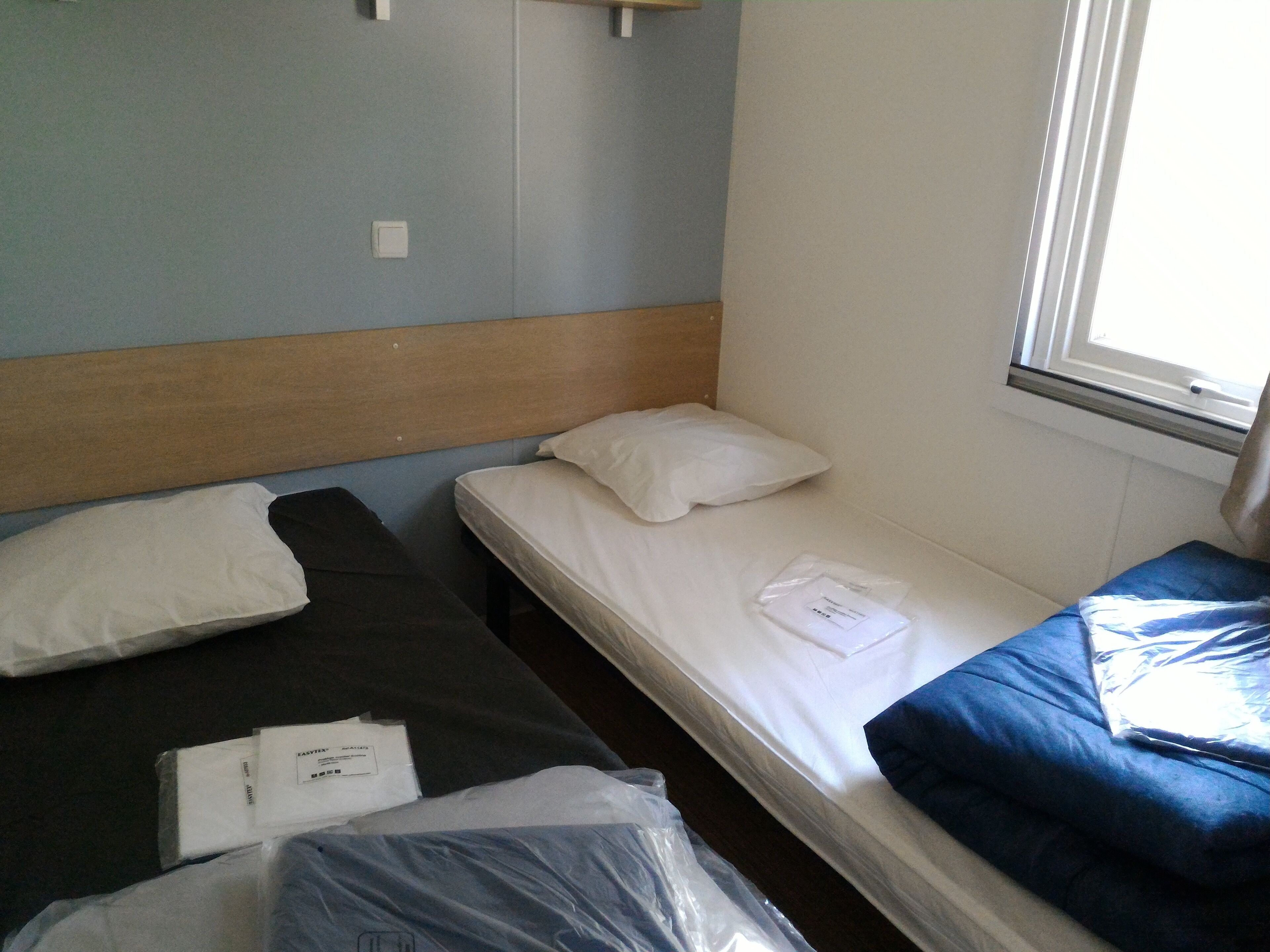 Une petite chambre avec un lit simple, une tête de lit en bois, une fenêtre et une vue sur l’extérieur.