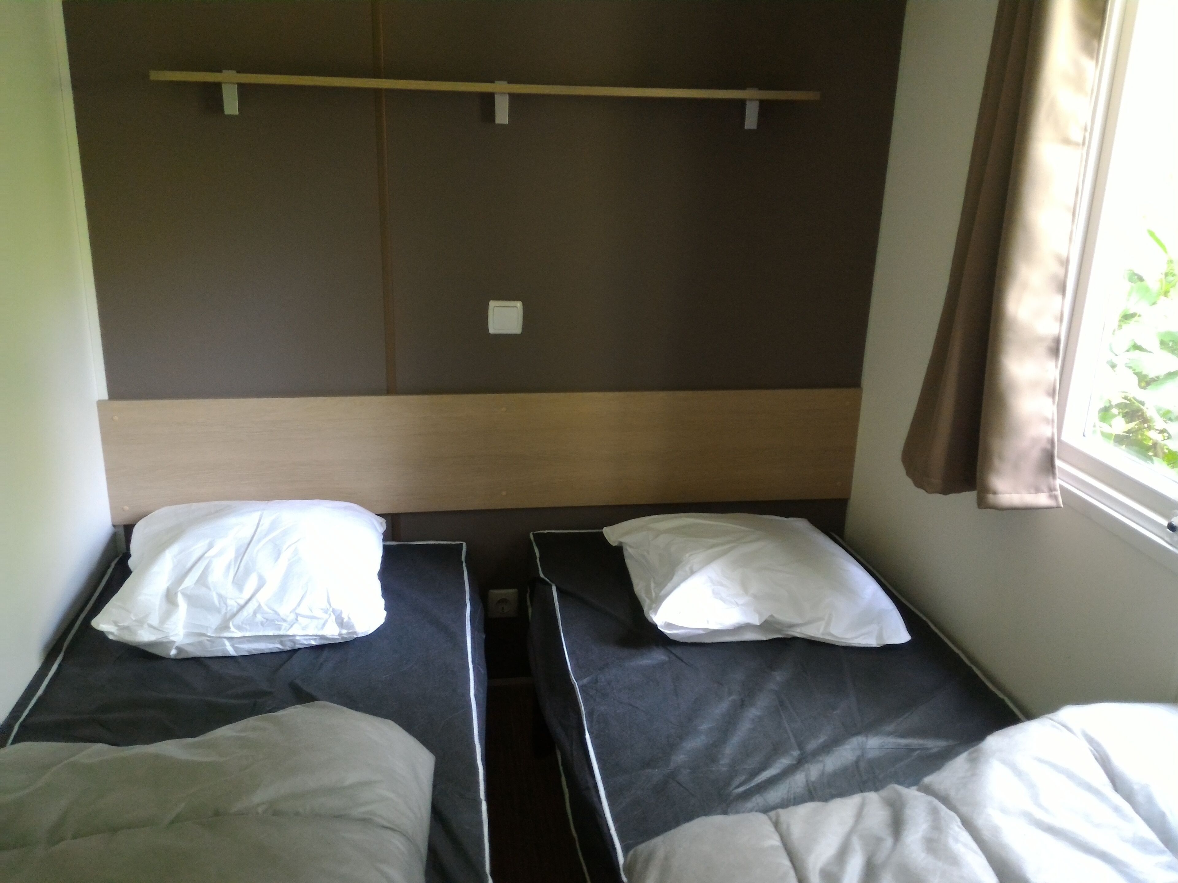 A room with two single beds, each with a pillow and a blanket. There is a wooden headboard, a window with curtains, and a wall-mounted light switch.