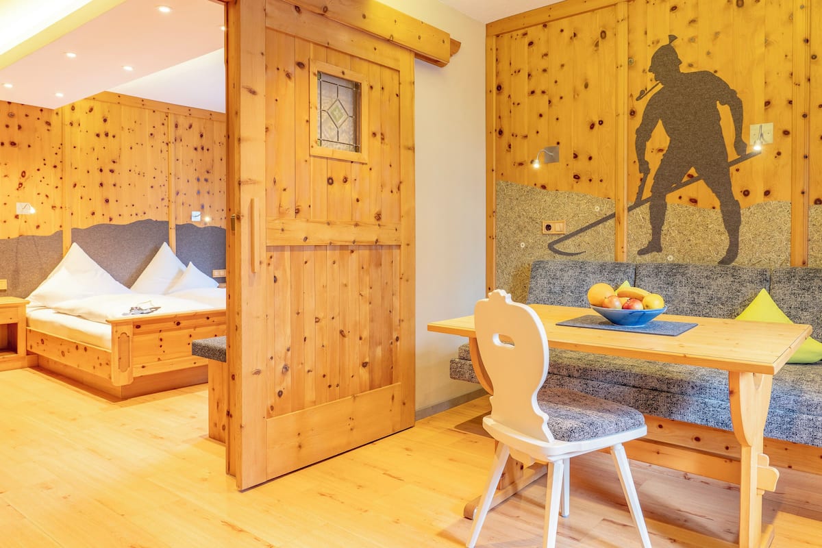 A wooden room with a bed, a table with a bowl of fruit, and a chair.