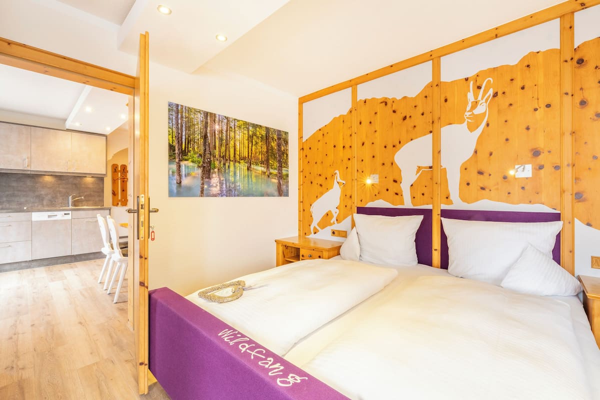 A modern bedroom with a wooden headboard, a painting of a forest, and a view of a kitchen with white cabinets and a dining area.