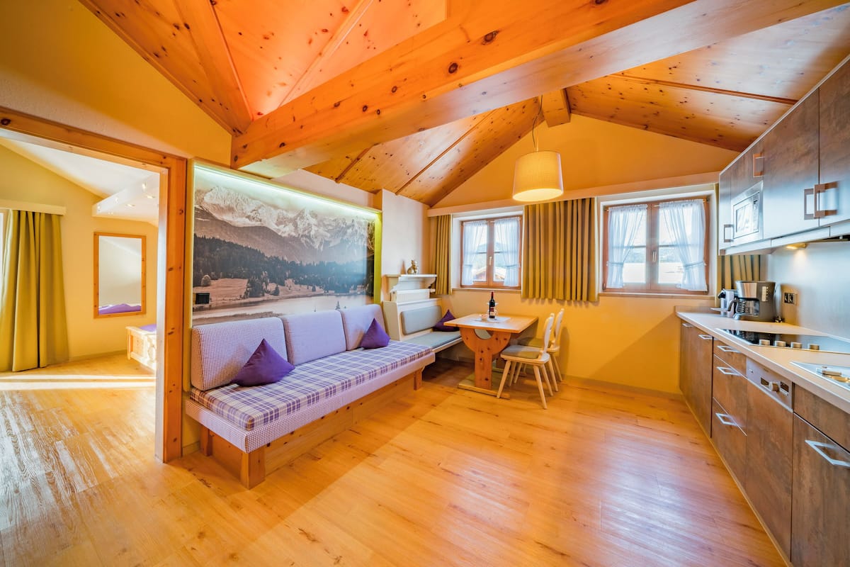 A cozy room with a wooden ceiling, a sofa with a checkered cover, a dining table with chairs, and a kitchen area with cabinets and a window.