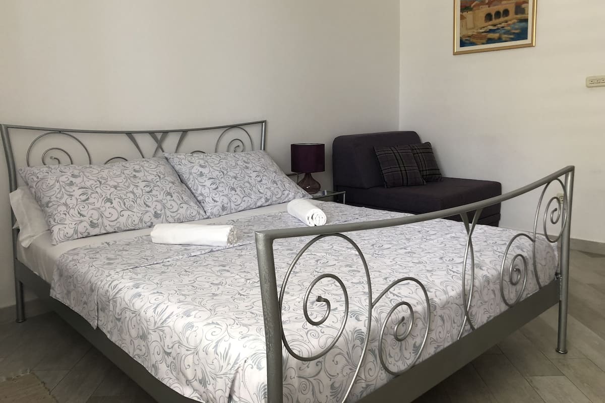 Uma cama bem arrumada com roupa de cama estampada, uma mesinha de cabeceira com abajur, um sofá escuro e um quadro na parede.