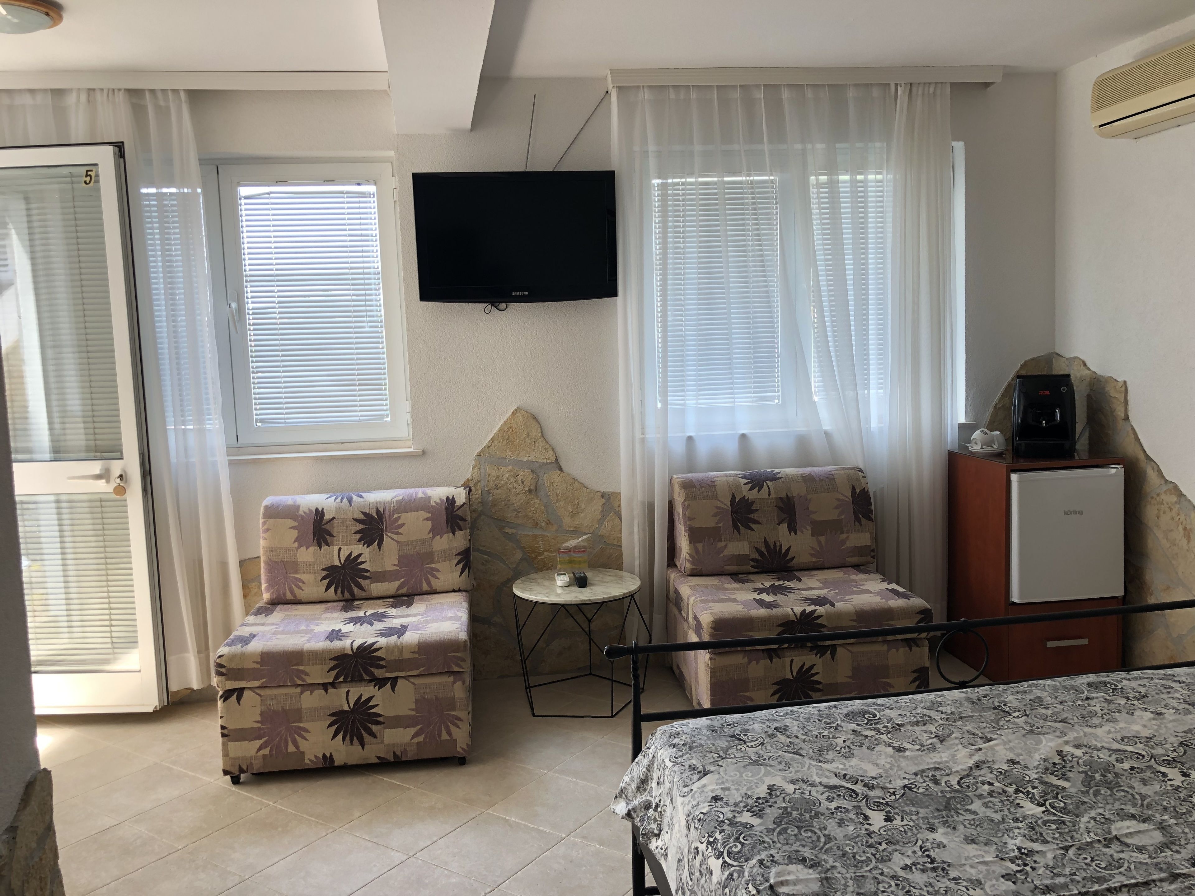 Una habitación con dos sillones estampados, una mesita, una cama con colcha estampada, un televisor y un refrigerador.