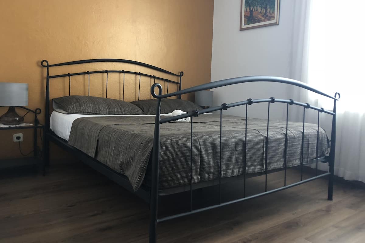 Uma cama de solteiro com estrutura de metal, uma mesa de cabeceira com luminária e um quadro na parede.