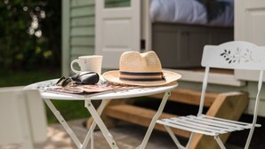 Ein kleiner runder Tisch mit einem Strohhut, Sonnenbrille und einer Tasse vor einem Haus.