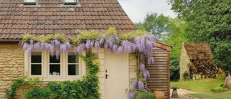 Une maison avec une façade en pierre, un toit en tuiles et un petit jardin agrémenté d’un banc et d’une plante en pot.