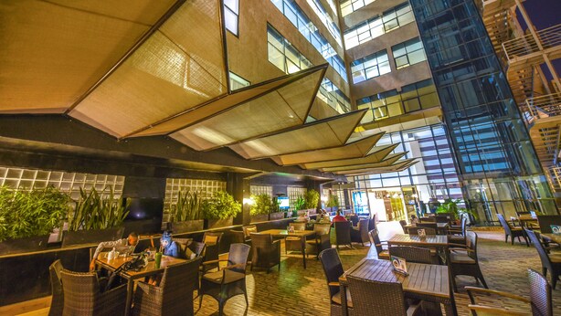 An outdoor dining area with tables and chairs, surrounded by large windows and plants. - BMK House Apartments (Kampala)