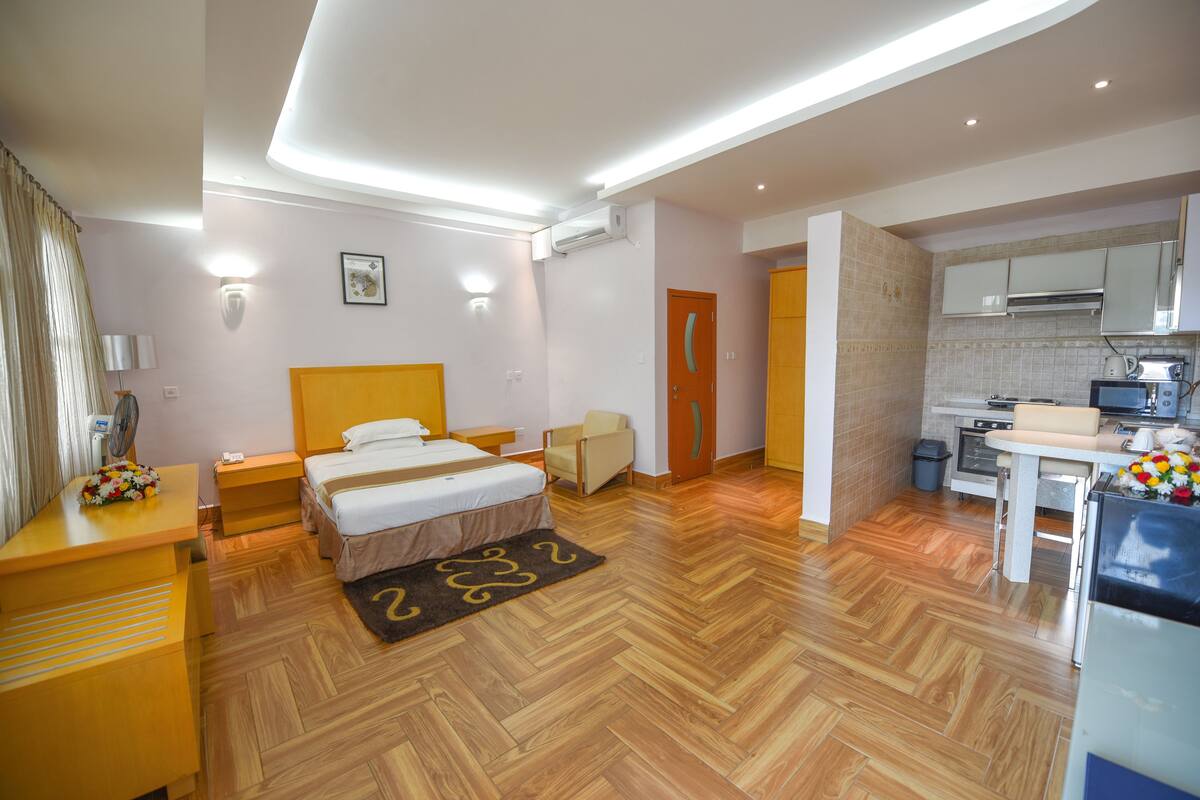 A modern hotel room with a bed, bedside table, chair, and a small kitchenette.