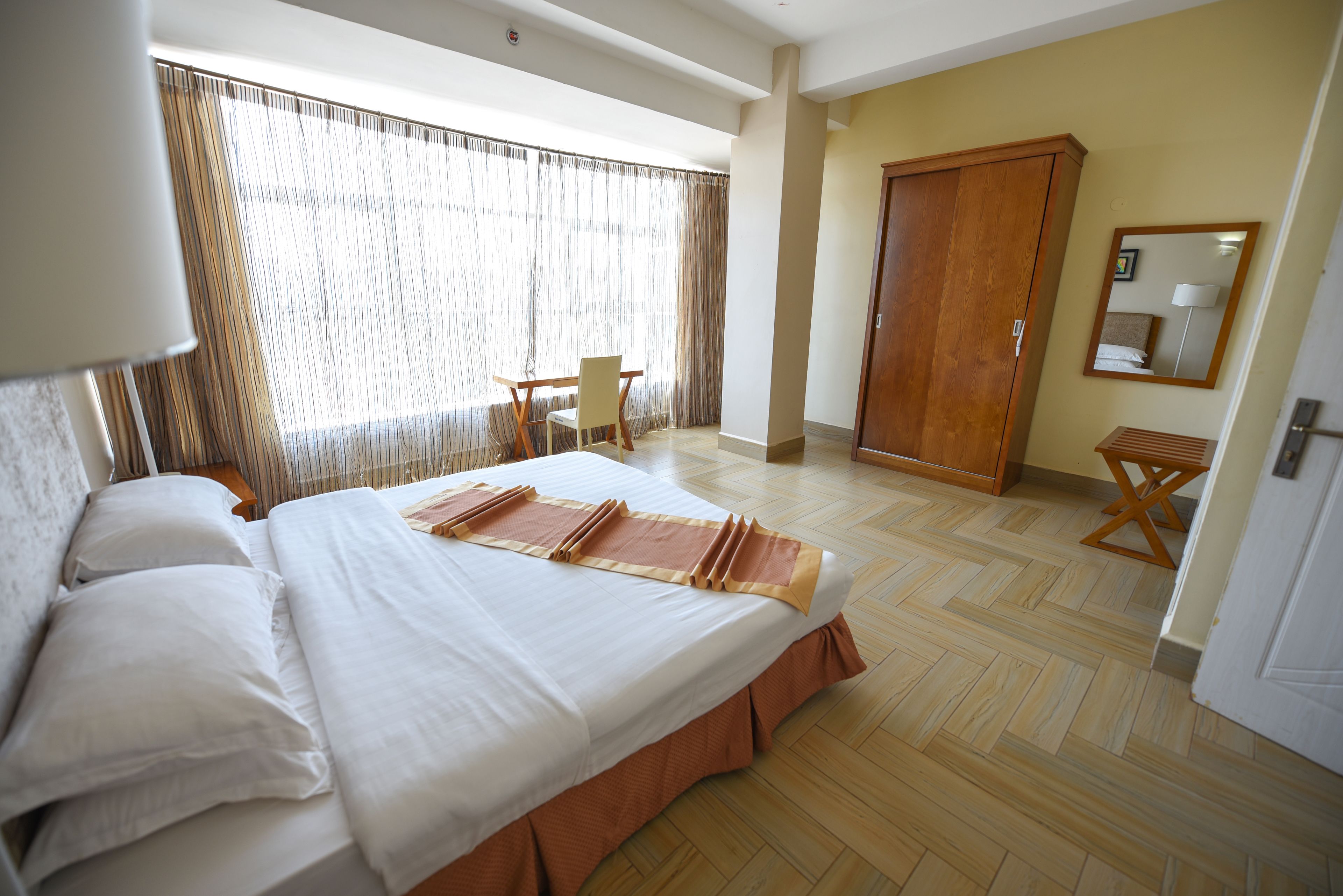 A hotel room with a large bed, a wooden wardrobe, a desk with a chair, and a window with curtains.
