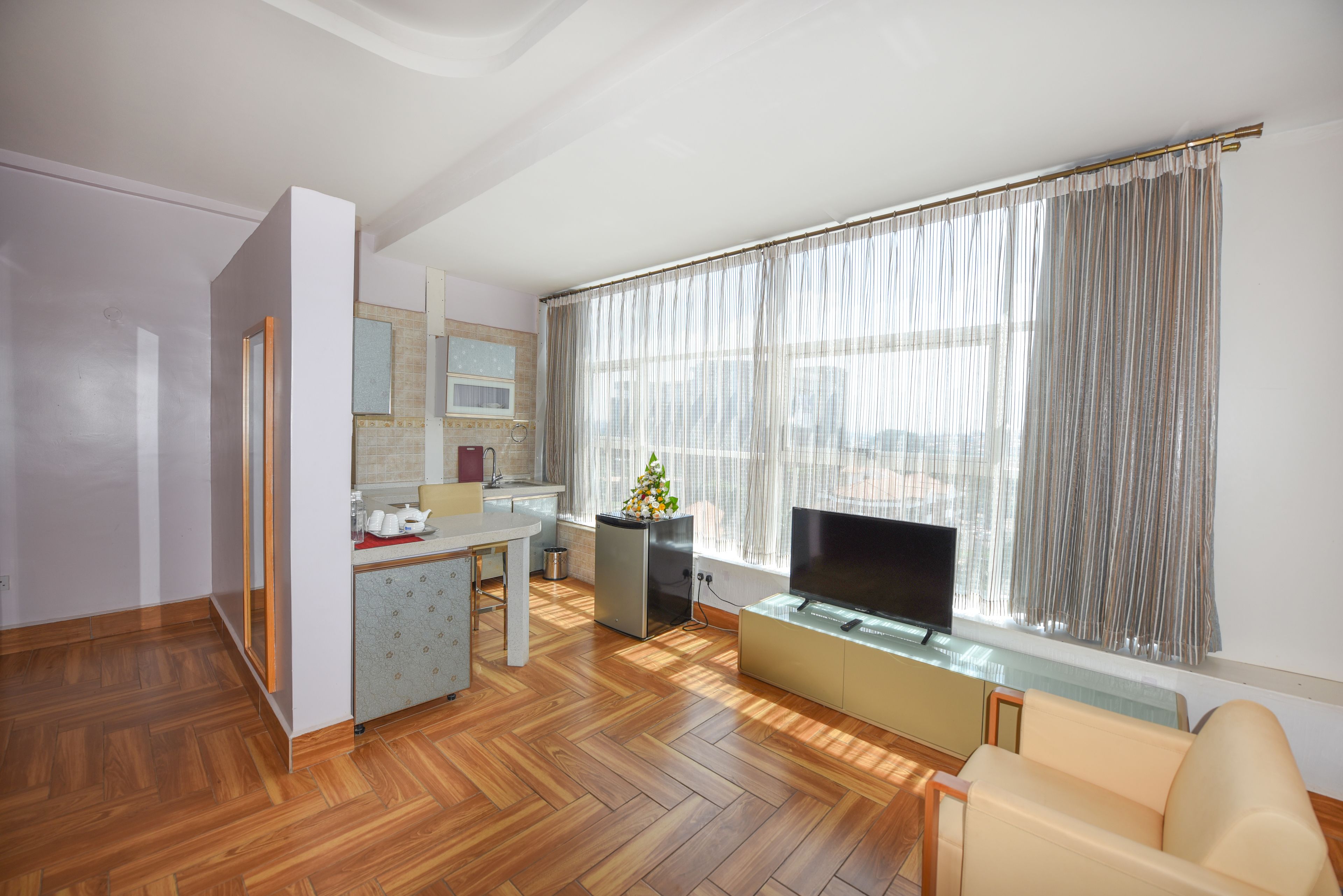 A modern living room with a wooden floor, a sofa, a TV on a glass stand, a kitchen area with a sink and cabinets, and a large window with curtains.