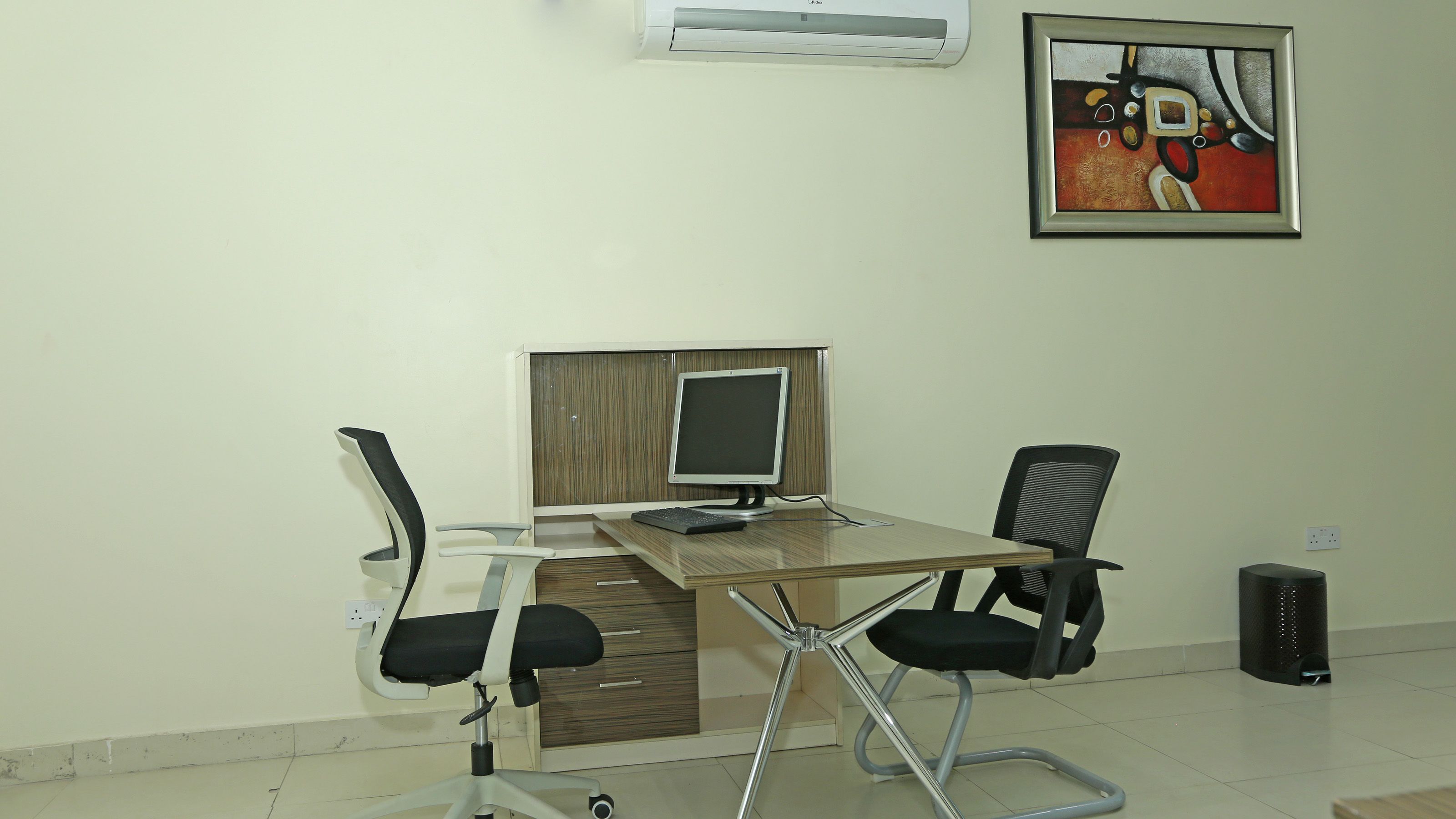 A modern office space with a wooden desk, two black office chairs, a computer monitor, and a framed abstract painting on the wall.