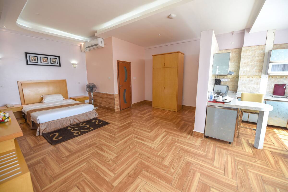 A hotel room with a bed, bedside table, fan, wardrobe, and kitchenette.