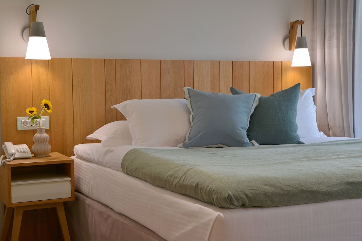 Een net opgemaakt bed met wit en groen beddengoed, een houten nachtkastje met een telefoon en een vaas zonnebloemen, en twee wandlampen.