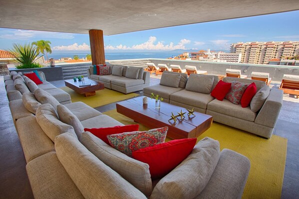 A rooftop lounge area with multiple sofas and a coffee table, overlooking a cityscape and the sea.