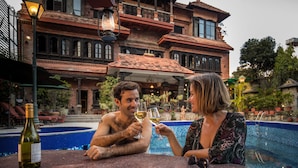 Ein Paar erhebt Weinkelche am Pool vor einem großen, verzierten Gebäude mit Ziegeldach.