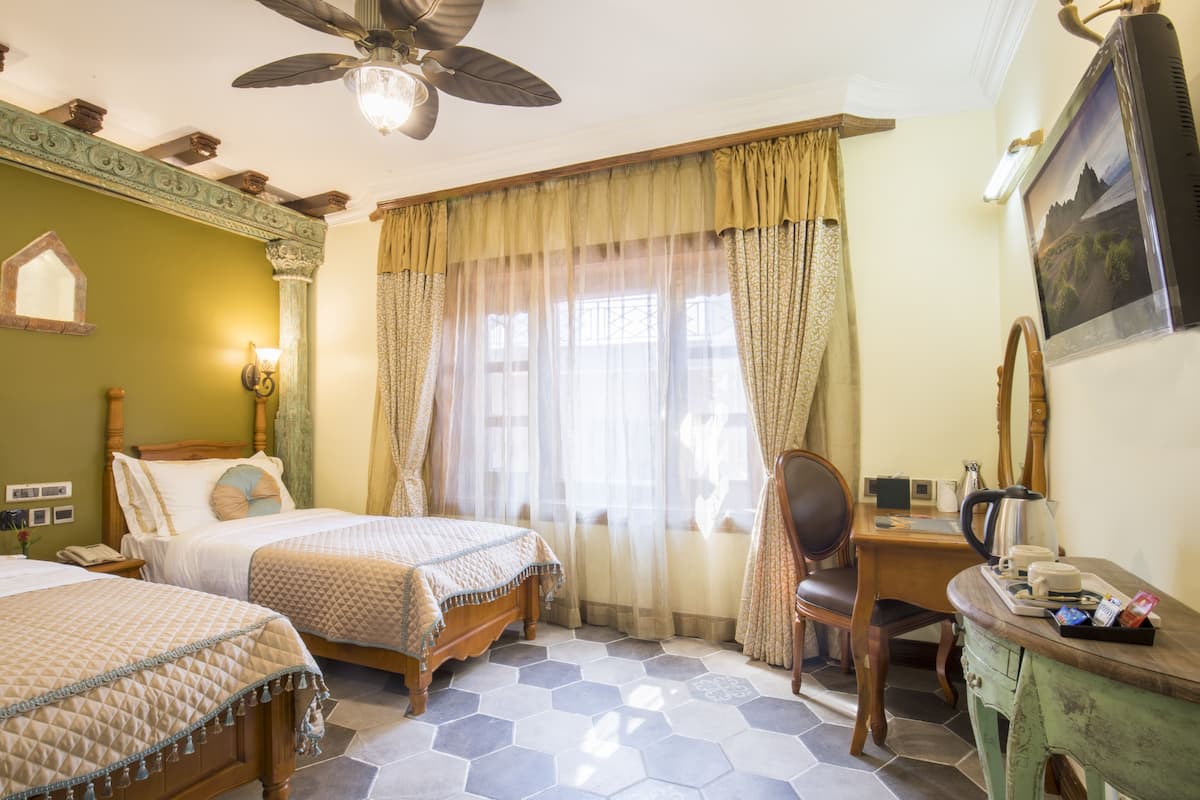 A hotel room with two beds, a desk, a chair, a ceiling fan, and a large window with curtains.