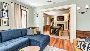 Ein Wohnzimmer mit blauem Sofa, Couchtisch und Kamin. An der Wand befinden sich Bilder in Rahmen, und im Hintergrund ist ein Essbereich zu sehen.