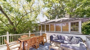 Eine Terrasse im Freien mit einem Holztisch und -stühlen, einem blauen Teppich mit Muster und einem violetten Sofa mit Kissen.