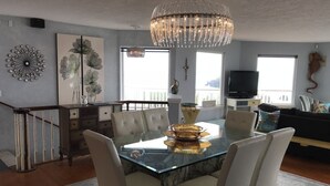 A dining area with a glass table, white chairs, a chandelier, artwork on the walls, and a staircase.