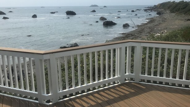 A wooden deck with white railings overlooking a rocky shoreline with the ocean in the background.